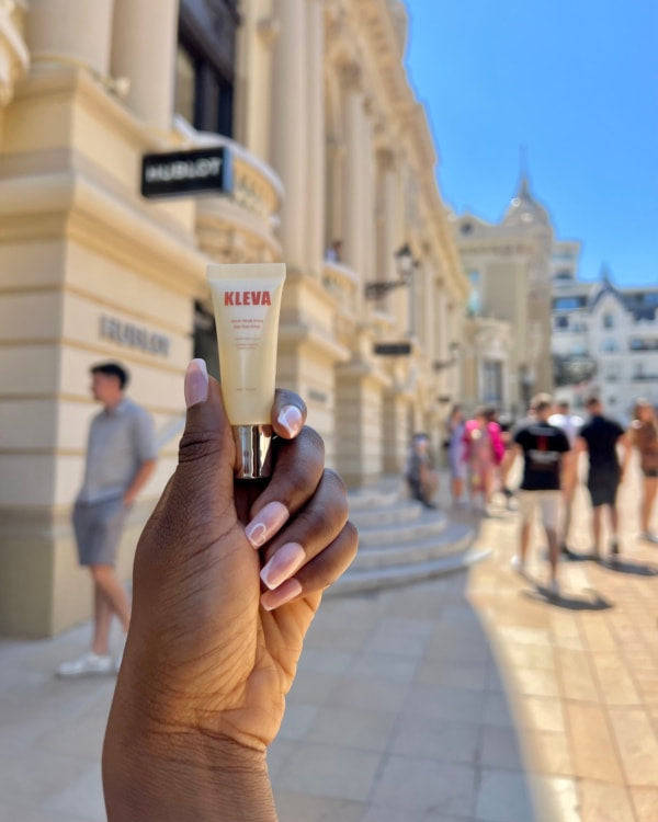 Hand holding a KLEVA body glue tube in front of luxury boutiques in Monaco under a sunny sky.