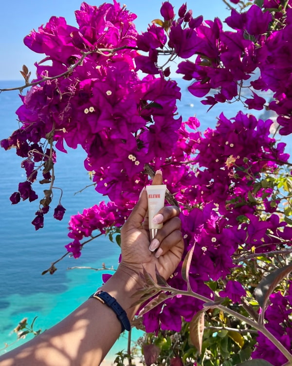 Main tenant un tube de colle corporelle KLEVA devant des fleurs de bougainvilliers roses éclatantes avec en arrière-plan la mer turquoise, évoquant l’usage en voyage et en été.